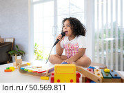 A tiny girl sits on a toy table, clutching a microphone, and sings cheerfully in the living room. Стоковое фото, агентство Ingram Publishing / Фотобанк Лори