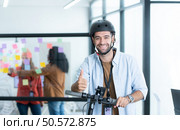 Portrait of smiling businessman riding electric scooter in office with colleague in background. Стоковое фото, агентство Ingram Publishing / Фотобанк Лори
