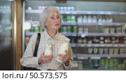 Mature woman with grocery cart standing near refrigerator with dairy products and choosing milk. Pensioner buys farm milk in an eco supermarket. Стоковое видео, видеограф Яков Филимонов / Фотобанк Лори
