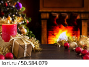 Christmas decorations, a gift and a candle in front of a fireplace. A fire is burning in the fireplace and Christmas stockings are hanging on the mantelpiece. A Christmas tree is standing next to the fireplace in the background. Стоковое фото, агентство Ingram Publishing / Фотобанк Лори