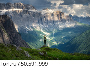 Sporty woman standing on a grassy hilltop with stunning view of the alpine mountains at sunset in summer. Scenery with girl, dramatic clouds over rocks and green valleys. Hiking in Dololmites, Italy. Стоковое фото, агентство Ingram Publishing / Фотобанк Лори