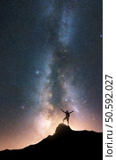 Milky Way and happy man with raised up arms on the mountain peak at starry night. Silhouette of guy, hill, sky with bright stars in summer. Galaxy. Space background. Vertical landscape with milky way. Стоковое фото, агентство Ingram Publishing / Фотобанк Лори