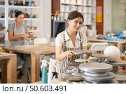 Girl in pottery workshop stands at table with tools, picks up rotate turntable revolving for work. Стоковое фото, фотограф Яков Филимонов / Фотобанк Лори