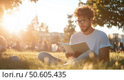 Young man enjoys reading a book in a sunlit park during a warm afternoon with friends in the background. Стоковое AI-изображение, AI-художник Сергей Толмачёв / Фотобанк Лори