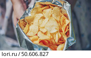 Woman grasping crinkled foil potato chip bag, displaying golden crispy snacks ready for casual outdoor munching. Стоковое фото, фотограф Mikhail Davidovich / Фотобанк Лори
