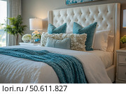 An elegant and cozy bedroom in a modern style. In the center of the frame is a luxurious bed with a high soft headboard, decorated with carefully placed decorative pillows in blue and white tones. A blanket in the same color as the pillows creates a feeling of coziness and comfort. The interior is complemented by bedside tables with lamps, fresh flowers and stylish details. Natural lighting and a calm color palette emphasize the atmosphere of coziness, cleanliness and modern design. An ideal photo for the theme of interior, decor and home comfort. Стоковое AI-изображение, AI-художник Анна Кривицкая / Фотобанк Лори