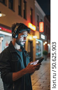 Hispanic young attractive man stands in dark street in front of shop, changes songs and tracks on smartphone, listens to music in wireless headphones. Hipster wearing casual dress. Стоковое AI-видео, аниматор Sergey Grishin / Фотобанк Лори