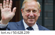 A man in a suit raises his hand in a greeting gesture, standing outdoors in front of a building entrance. Стоковое AI-видео, аниматор Sergey Grishin / Фотобанк Лори