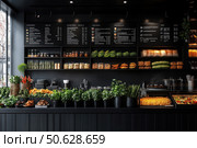 A modern interior of a cafe or restaurant with an open kitchen and eco-minimalist design. Dark matte surfaces highlight the bright colors of fresh vegetables, herbs and spices, neatly arranged on the counter and shelves. In the background is a stylish black menu on the wall with white inscriptions. The interior is dominated by natural elements: greenery in pots, wicker baskets, glass jars with nuts, spices and pickles. The atmosphere of the place is clean, modern and appetizing, ideal for presenting a gastronomic brand, healthy food or a farm-to-table concept. Стоковое AI-изображение, AI-художник Анна Кривицкая / Фотобанк Лори