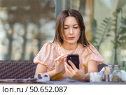 Молодая красивая европейка с длинными волосами держит в руках смартфон, проверяет социальные сети, пользуется мобильным приложением, сидя за столиком на открытом воздухе в уютном современном кафе. Стоковое фото, фотограф Роман Дорохов / Фотобанк Лори