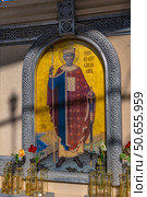 The photograph depicts an icon of the Holy Prince Vladimir, Equal to the Apostles, installed in a carved stone icon case on the territory of the New Chersonesos historical and cultural complex in Sevastopol. The holy prince is depicted in royal vestments with a cross and a sword, on a gold background, symbolizing holiness. Glass vases with live roses are installed in front of the icon, emphasizing the veneration and solemnity of the place. The Byzantine ornament of the frame emphasizes the sacred nature of the composition. Стоковое фото, фотограф Анна Кривицкая / Фотобанк Лори