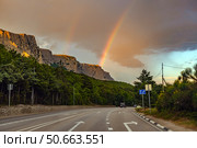 Picturesque landscape of the southern coast of Crimea at sunset. The asphalt road leads along a dense green forest to a rock massif, illuminated by the soft orange light of the setting sun. Two rainbows spread out over the mountains, one of which stands out especially brightly against the background of clouds. The unique combination of natural phenomena - sunset light, dramatic clouds and a double rainbow - creates an atmosphere of magic and peace. In the frame - a road with markings, cars, a pine forest and majestic rocks, typical of Crimea. Стоковое фото, фотограф Анна Кривицкая / Фотобанк Лори