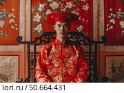 Groom wearing traditional red ao dai sitting on wooden chair. Стоковое AI-изображение, AI-художник Иван Рябоконь / Фотобанк Лори