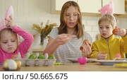 Children siblings painting Easter egg sitting together at table. Easter Family traditions. Teen girl teaching happy little brother and sister to dye, decorate eggs with paints. Preparation to holidays. Стоковое видео, видеограф Марина Демидюк / Фотобанк Лори