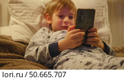 Cute kid boy in pajamas lying in bed watching smartphone, smiling. Children's screen time. Child plays games using phone at the evening or night. Social media addiction. Childhood, modern technology. Стоковое видео, видеограф Марина Демидюк / Фотобанк Лори