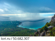 Stunning view of the Crimean coast from above. In the foreground you can see a rocky area covered with dense green vegetation, smoothly descending to the shore of the Black Sea. In the background are small buildings and residential complexes located along the coastal zone.The most impressive detail of the photo is the rain front hanging over the sea. The boundary between the clear and rainy zone is clearly visible, where streams of rain descend from the clouds and create the effect of a blurred horizon. This spectacle emphasizes the unique microclimate of Crimea, where mountains, sea and clouds form unpredictable weather phenomena. (2021 год). Стоковое фото, фотограф Анна Кривицкая / Фотобанк Лори