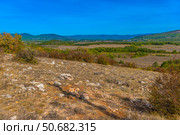 Panoramic view of a dry rocky landscape in Crimea with sparse vegetation, low bushes and boulders in the foreground. In the distance are green forest belts, fields and mountain ranges under a bright blue sky. The typical southern landscape conveys an atmosphere of peace, solitude and natural pristineness. Ideal for topics related to ecotourism, geography, nature and Crimea. Стоковое фото, фотограф Анна Кривицкая / Фотобанк Лори