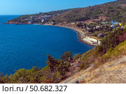 A magnificent view of the Crimean coast. From above, a panorama of the curved coastline, covered with sparse vegetation, rocks and junipers, opens up. In the distance, a small town or village with white houses spread out along the slope of a hill is visible. The Black Sea is deep blue, calm, with slight glare from the sun. On the horizon, closer to the center of the frame, an old sea platform or pier is noticeable. This place conveys an atmosphere of privacy, tranquility and southern warmth. Стоковое фото, фотограф Анна Кривицкая / Фотобанк Лори