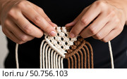Close-up of hands weaving macrame with brown and white cords. The person has light skin and is focused on the intricate knotting process. Стоковое AI-изображение, AI-художник Журавлева Кристина / Фотобанк Лори