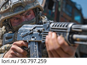 Российский военнослужащий в полной боевой экипировке и защитном шлеме прицеливается из автомата на фоне военной техники в зоне боевых действий. Стоковое AI-изображение, AI-художник ArtVladZ / Фотобанк Лори