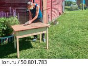 Grinding wooden tabletop with angle grinder, handyman wearing respirator and safety glasses working outdoors in rural environment. Стоковое фото, фотограф Владимир Григорьев / Фотобанк Лори