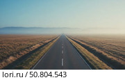 A long, straight road stretches through a vast, open landscape. The horizon is clear with a blue sky and distant mountains. Fog covers the ground, creating a serene atmosphere. Стоковое AI-видео, аниматор Olga Dmitrieva / Фотобанк Лори