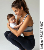 A young mother in a tracksuit holds her newborn baby in her arms. She does fitness exercises with her child. Стоковое AI-изображение, AI-художник Ирина Гильмуллина / Фотобанк Лори