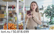 Carefree young woman in thirties engrossed in video call on smartphone while shopping in local grocery store, holding basket full of purchases. Стоковое видео, видеограф Яков Филимонов / Фотобанк Лори