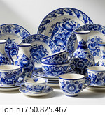 Blue and white ceramic tableware features intricate floral patterns, birds, and fruits, including plates, cups, vases, and saucers, all artistically arranged in an elegant display. Стоковое AI-изображение, AI-художник Olga Dmitrieva / Фотобанк Лори