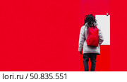 Back View: Person with Dreadlocks and Red Backpack, standing near white space against a vibrant red and white panel. Стоковое AI-изображение, AI-художник Данила Большаков / Фотобанк Лори