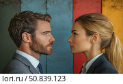 Business portrait of a man and a woman facing each other in a tense confrontation. They are dressed in formal suits, which emphasizes the business context, and the background of colored wooden panels adds artistic expressiveness. The image symbolizes rivalry, leadership, gender equality, negotiations and the power of the gaze. Suitable for themes: gender relations, business communication, equality, corporate ethics. Стоковое AI-изображение, AI-художник Анна Кривицкая / Фотобанк Лори