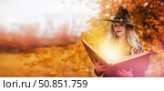 Halloween a woman with a magic spell book in a witch hat is conjuring, performing witchcraft over the autumn background of the forest. Стоковое фото, фотограф Лазаренко Светлана / Фотобанк Лори