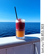 A bright double-layered cocktail and a black straw on the deck against the background of the deep blue sea and clear sky. The atmosphere of summer relaxation and freedom. Yacht vacation. Стоковое фото, фотограф Анна Чижова / Фотобанк Лори