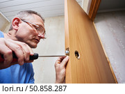 Installer door fastens metal latch plate to wooden door edge using screwdriver during handle installation process. Стоковое фото, фотограф Владимир Григорьев / Фотобанк Лори