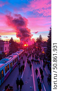 Street Riot: A vivid urban scene, people filling the roadway with a tram, and a fiery smoke plume under a dramatically colored sky. Стоковое AI-изображение, AI-художник Данила Большаков / Фотобанк Лори