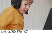 Little smiling 6 years old boy using a laptop with headphones. Child having video call with friends, granny at home in bed. Kid looking at display and waving hand. Childhood leisure activity concept. Стоковое видео, видеограф Марина Демидюк / Фотобанк Лори