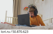 Little smiling 6 years old boy using a laptop with headphones. Child having video call with friends, granny at home in bed. Kid looking at display and waving hand. Childhood leisure activity concept. Стоковое видео, видеограф Марина Демидюк / Фотобанк Лори