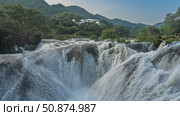 A stormy mountain river. Streams of white water are bubbling and foaming on boulders in the riverbed. Green vegetation on the shores. A building on a hill. China. Silver chain falls. Guizhou. Стоковое фото, фотограф Вера Ивановна Кубарева / Фотобанк Лори