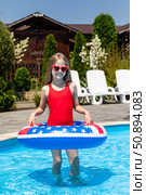 Happy little girl in red swimsuit and sunglasses stands in outdoor swimming pool, has fun and dances with a lifebuoy decorated with American flag print. Vacation in USA concept. 4th of July pool party. Стоковое фото, фотограф Ирина Михайличенко / Фотобанк Лори
