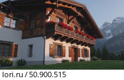 Rustic wooden Swiss chalet glowing under late afternoon sun, traditional balconies decorated with bright red geraniums, nestled before dramatic alpine mountain backdrop. Стоковое AI-видео, аниматор Данил Руденко / Фотобанк Лори