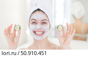 Cheerful woman with a towel on her hair and in a clay face mask fooling around with cucumbers in her hands. Taking care of beauty at home. Стоковое фото, фотограф Михаил Решетников / Фотобанк Лори