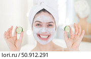 Cheerful woman with a towel on her hair and in a clay face mask fooling around with cucumbers in her hands. Taking care of beauty at home. Стоковое фото, фотограф Михаил Решетников / Фотобанк Лори