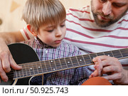 Handsome dad teaches his cute son to play the guitar sitting on sofa in the living room at home. Happy Father's Day. Father teaching his child to play guitar. Kid Learning music skill. Art education. Стоковое фото, фотограф Марина Демидюк / Фотобанк Лори