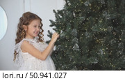 Happy beautiful little girl in elegant white fluffy dress stands near a Christmas tree at home, laughing joyfully in anticipation of winter holiday. New year, fairy tales and children dreams footage. Стоковое видео, видеограф Ирина Михайличенко / Фотобанк Лори