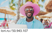 Concept: Beach office. Portrait of an African-American man with laptop, pink sunhat, and sunglasses, smiling on a tropical beach. Стоковое AI-изображение, AI-художник Данила Большаков / Фотобанк Лори