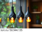 Modern pendant lamps with matte black shades and warm yellow light in an interior with elements of wood and greenery. The laconic and elegant design makes these lamps ideal for cafes, restaurants, living rooms or offices in loft style, Scandinavian or minimalist design. Soft glow creates a cozy atmosphere and emphasizes the texture of the materials. Стоковое AI-изображение, AI-художник Анна Кривицкая / Фотобанк Лори