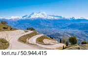 Scenic mountain road winding through valleys, with snow-capped peaks under a clear blue sky, offering a breathtaking view of nature. Стоковое AI-изображение, AI-художник Данила Большаков / Фотобанк Лори