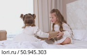Two sisters wearing white dresses, sitting on a bed with white bedding, pulling big wrapped gift box, fighting, cannot share a present on the holiday morning at home. Стоковое видео, видеограф Ирина Михайличенко / Фотобанк Лори
