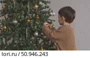 Side view of a cute little boy wearing a brown warm sweater, decorating a Christmas tree in the living room at home. Merry Christmas and happy New Year winter holidays. High-quality footage. Стоковое видео, видеограф Ирина Михайличенко / Фотобанк Лори