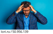 Stressed adult male with glasses holding his head, depicting a headache, mental strain, or overwhelm against a blue solid wall. Стоковое AI-изображение, AI-художник Данила Большаков / Фотобанк Лори